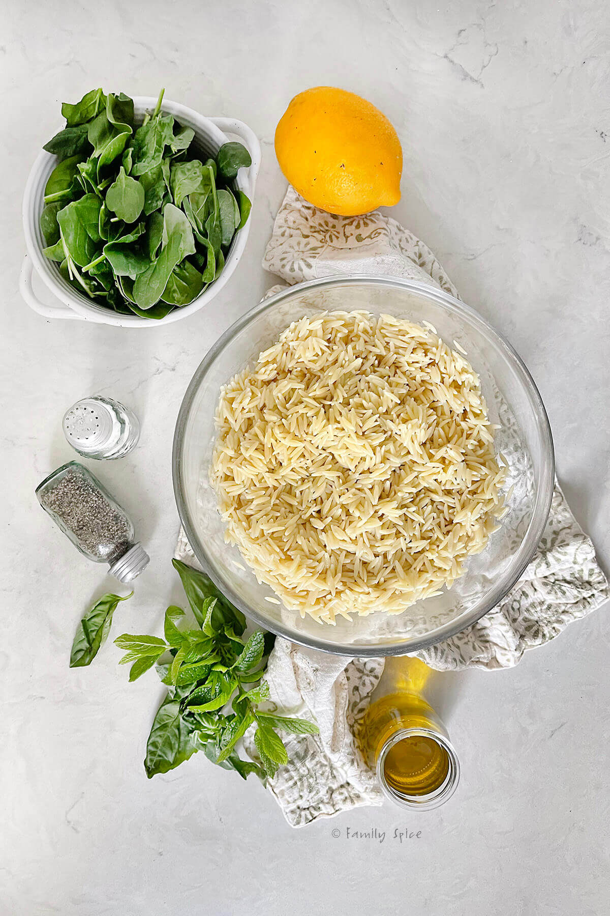 A glass mixing bowl with cooked orzo pasta in it with a bowl of fresh spinach next to it, along with a lemon, salt and pepper, fresh herbs and olive oil