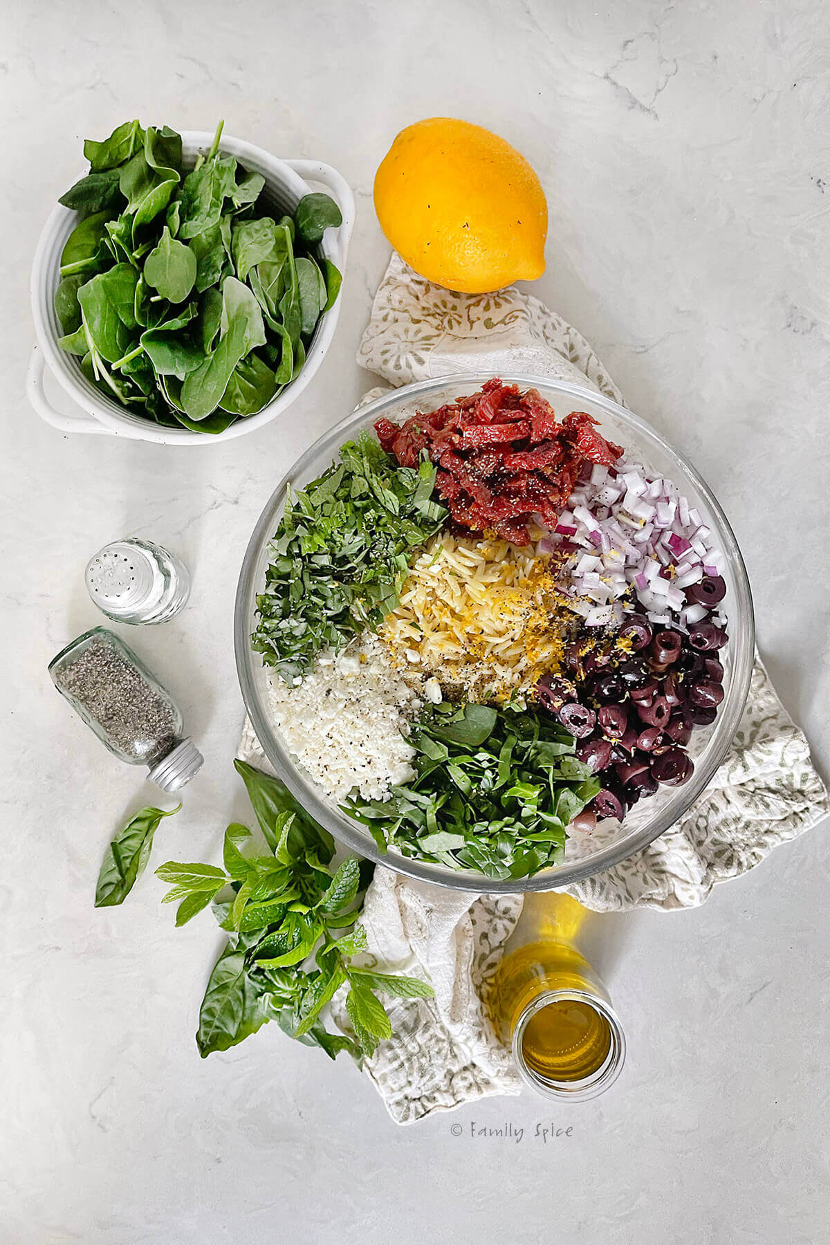A glass mixing bowl with orzo pasta, chopped spinach, herbs, feta, olives, red onions and sun dried tomatoes in it with a bowl of fresh spinach next to it, along with a lemon, salt and pepper, fresh herbs and olive oil