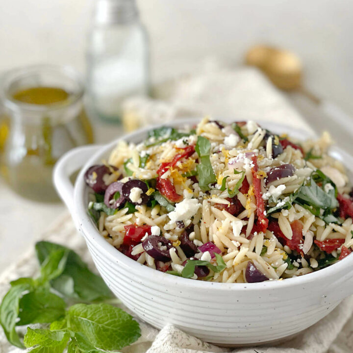 Side view of a white bowl with orzo pasta salad in it with fresh mint next to it and olive oil, salt and pepper behind it