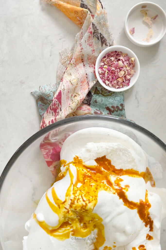 Top view of a large glass mixing bowl with melty vanilla ice cream topped with saffron water and other ingredients next to it