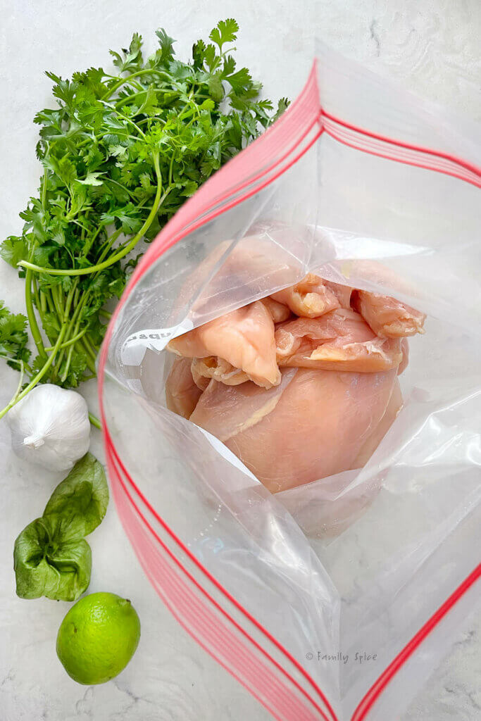 A large resealable bag with raw chicken breast in it and cilantro, garlic and lime next to it