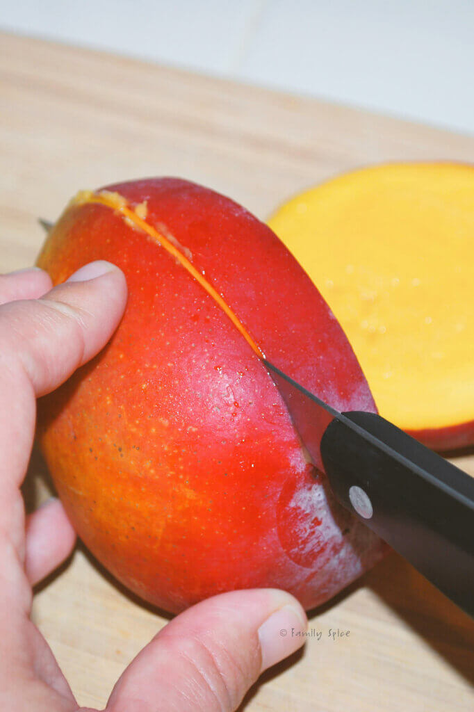 A hand holding a halved mango and knife cutting out the pit
