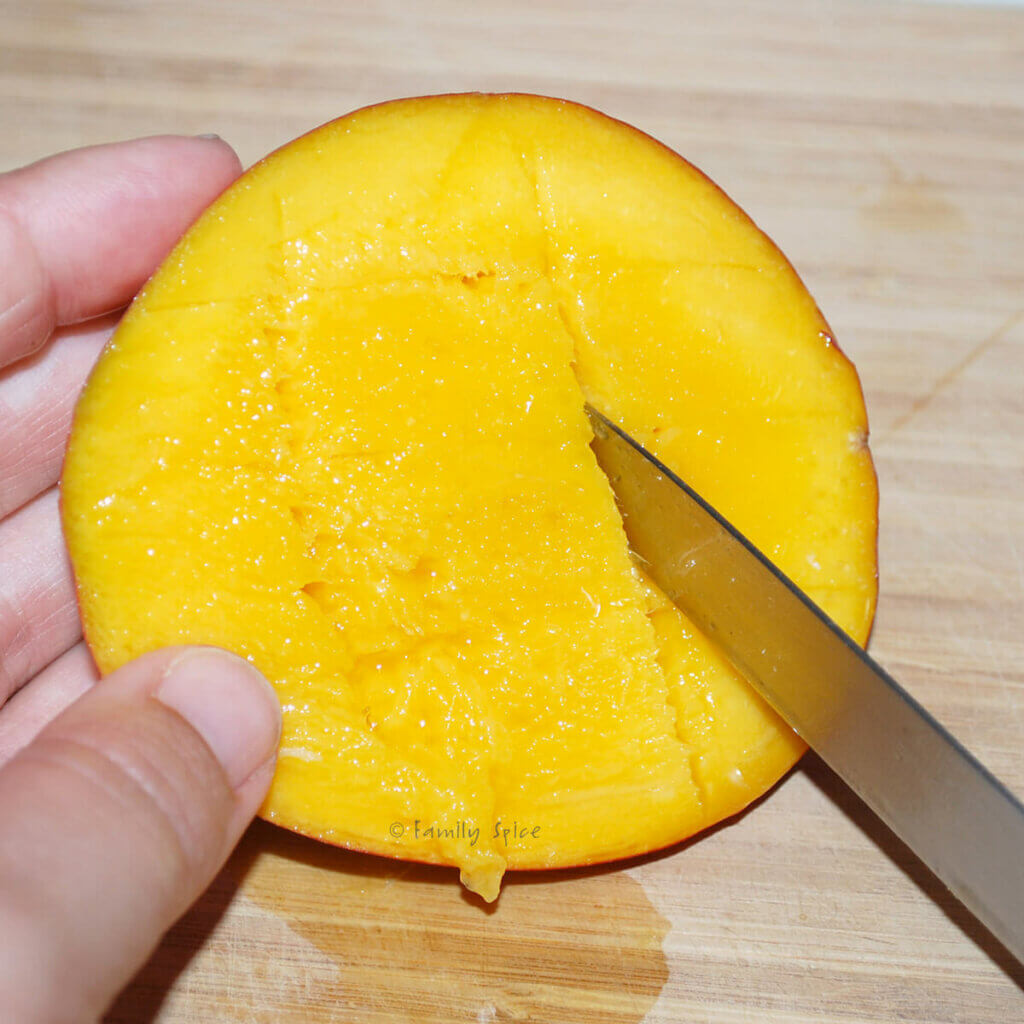 A hand holding a mango half with a knife scoring cubes in it