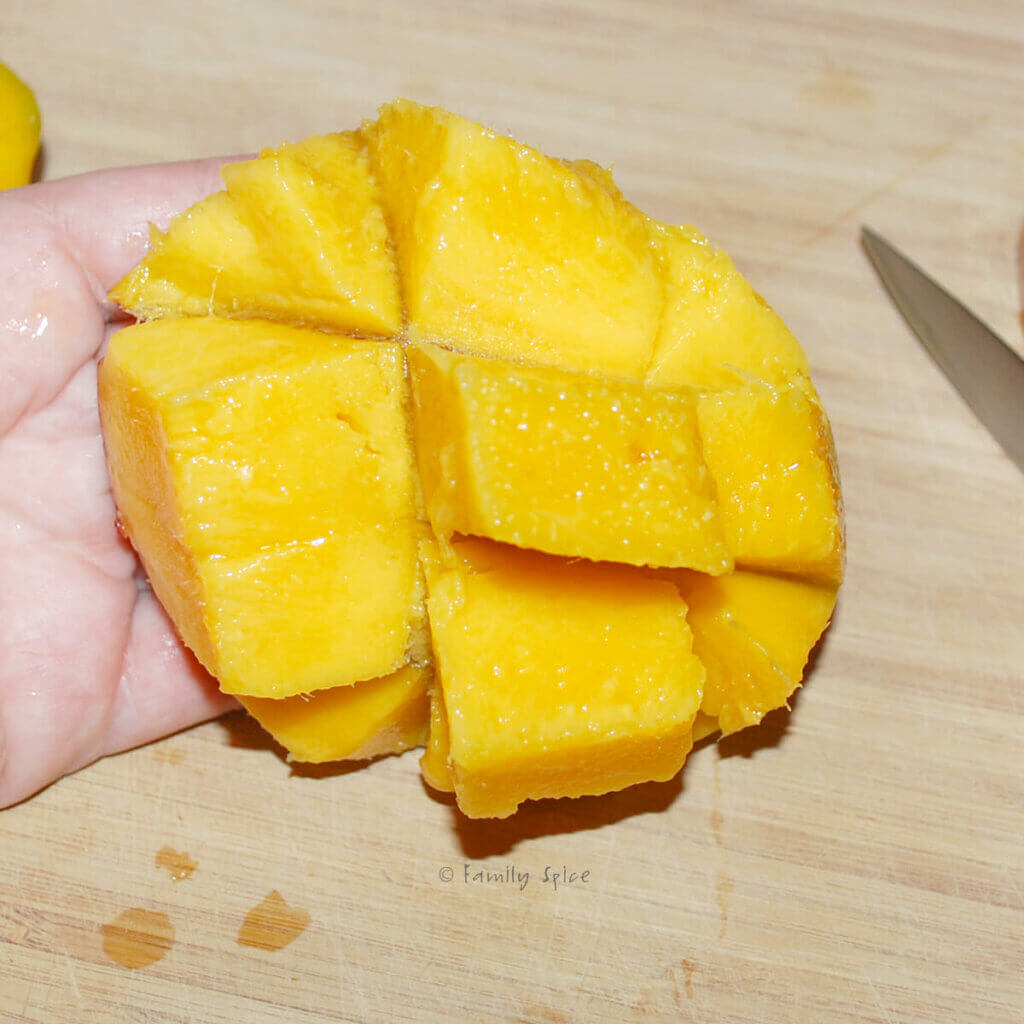 A hand holding a mango half with cubes scored in it and inverted
