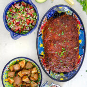 A Mexican serving dish with chorizo meatloaf loaf on it, a small bowl with Shirazi salad, another with roasted seasoned potatoes and another with chopped radishes