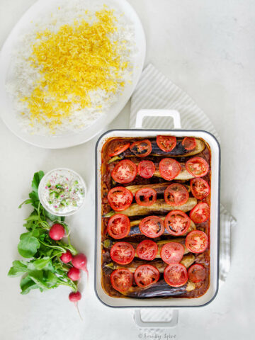 A rectangular pan with baked Perisan eggplant stew (khoresh bademjoon) a platter of Persian rice, a bowl of mast o khiar and fresh radishes