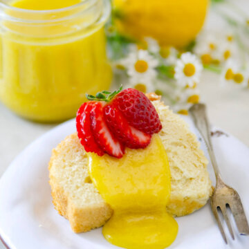 Closeup of a slice of sponge cake topped with lemon curd and strawberries with a jar of curd behind it.