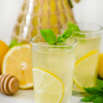 Closeup of a glass with honey lemonade in it and pitcher of lemonade and lemons behind them.