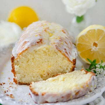 Side view of a glazed lemon tea bread on a white plate with lemons and white flowers around it.
