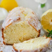 Closeup of a glazed lemon tea bread on a white plate with lemons and white flowers around it.