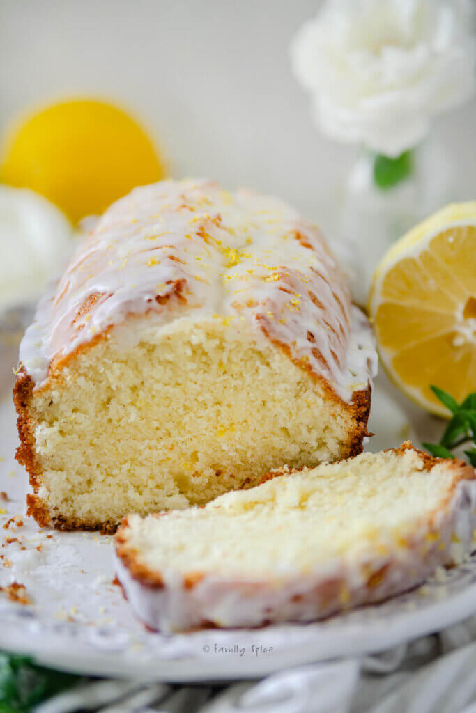 Closeup of a glazed lemon tea bread on a white plate with lemons and white flowers around it.