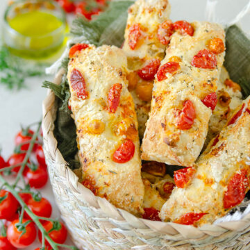 Side view of a basket with savory tomato scones in it with cherry tomatoes, olive oil and rosemary around it.