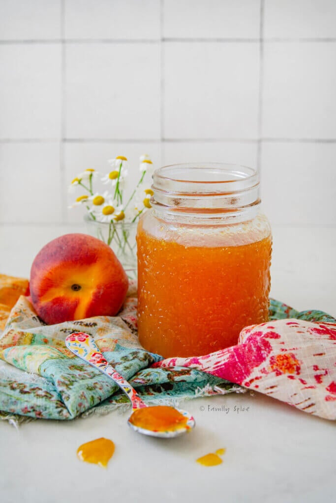 Closeup of a bottle of peach jam on a pretty napkin with a peach next to it.