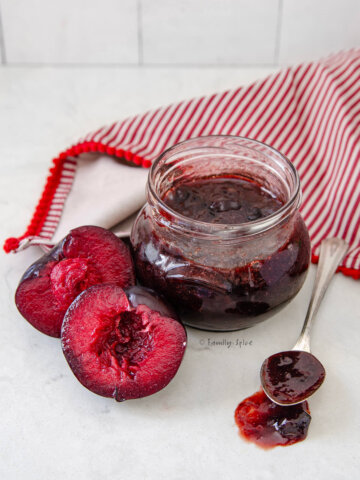 A jar of plum jam with a spoon of jam and more plums next to it.