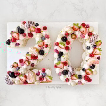 A number 20 birthday cake decorated with white frosting, fruit and assorted candies and flowers on a white background.
