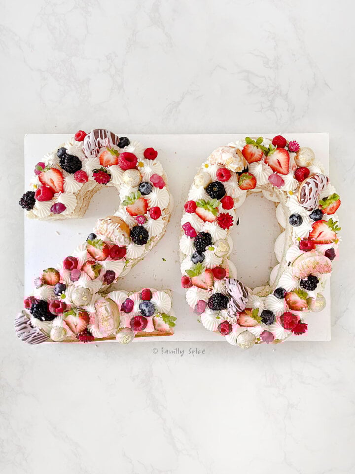 A number 20 birthday cake decorated with white frosting, fruit and assorted candies and flowers on a white background.