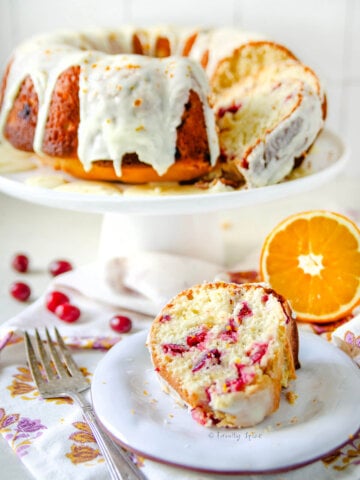 A slice of orange cranberry olive oil cake on a plate with the rest of the bundt cake behind it.