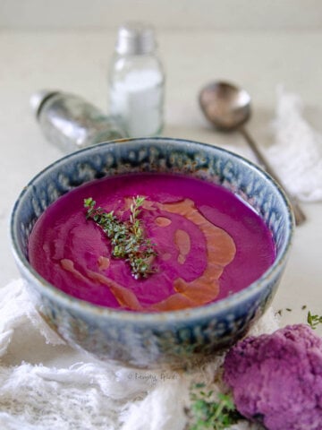 Side view of a bowl of purple sweet potato soup with fresh thyme on a white marble countertop.