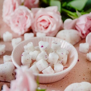 Closeup of a small pink bowl filled with heart shaped sugar cubes on a pink background surrounded by more sugar cubes and pink roses.