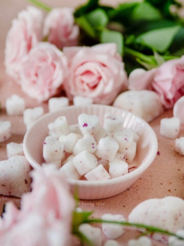 Closeup of a small pink bowl filled with heart shaped sugar cubes on a pink background surrounded by more sugar cubes and pink roses.