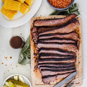 Top view of an oven baked brisket sliced up on a cutting board with corn, beans and pickle around it.
