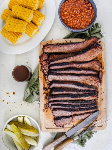 Top view of an oven baked brisket sliced up on a cutting board with corn, beans and pickle around it.