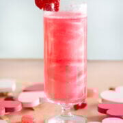 Side view of a short stemmed champagne flute filled with shimmering pink mimosa garnished with a heart shaped strawberry.