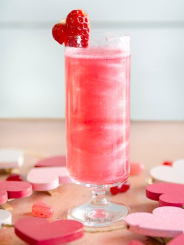 Side view of a short stemmed champagne flute filled with shimmering pink mimosa garnished with a heart shaped strawberry.