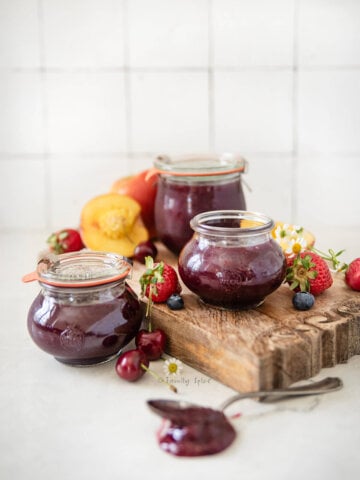 Three jars of mixed fruit jam on a cutting board with fruit around it.