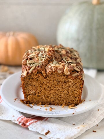 Closeup of a loaf of olive oil pumpkin bread sliced open and two pumpkins behind it.